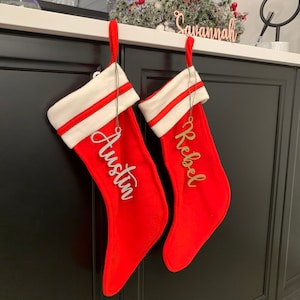 May include: Two red Christmas stockings with white trim and personalized name tags. The stockings are hanging on a black shelf with the name "Savannah" on a wooden sign above them. The name tags are made of wood and are in a cursive font. The stocking on the left has the name "Austin" on it, and the stocking on the right has the name "Reba" on it.