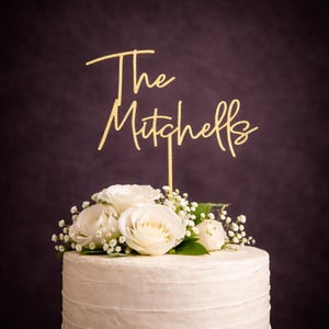 May include: A gold glitter cake topper featuring the text The Mitchells in a cursive script font, positioned atop a white frosted cake decorated with a cluster of white roses and small white baby's breath flowers against a dark background.