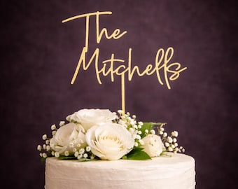 May include: A gold glitter cake topper featuring the text The Mitchells in a cursive script font, positioned atop a white frosted cake decorated with a cluster of white roses and small white baby's breath flowers against a dark background.