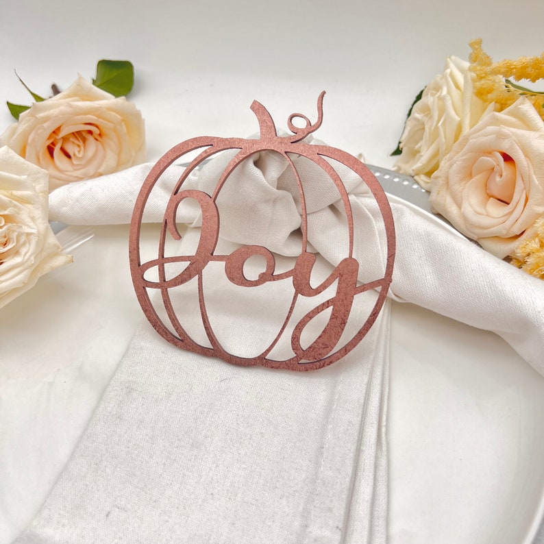May include: A wooden pumpkin-shaped napkin ring with the word "Joy" cut out in the centre. The napkin ring is a light brown colour and is sitting on a white napkin.