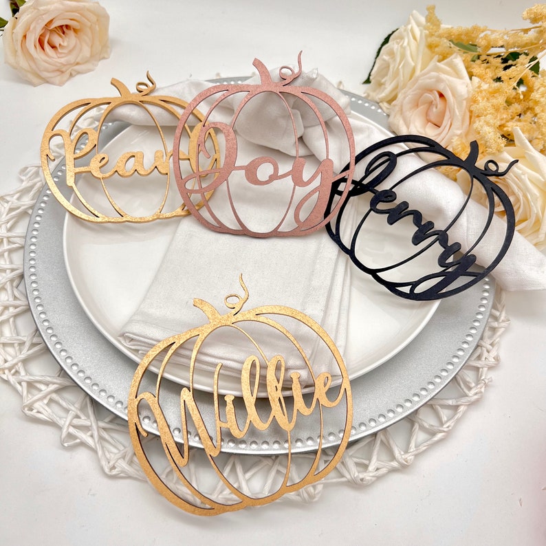 May include: Four wooden pumpkin place card holders with the names "Pear", "Joy", "Jenny", and "Willie" written in cursive. The place cards are in gold, rose gold, and black.