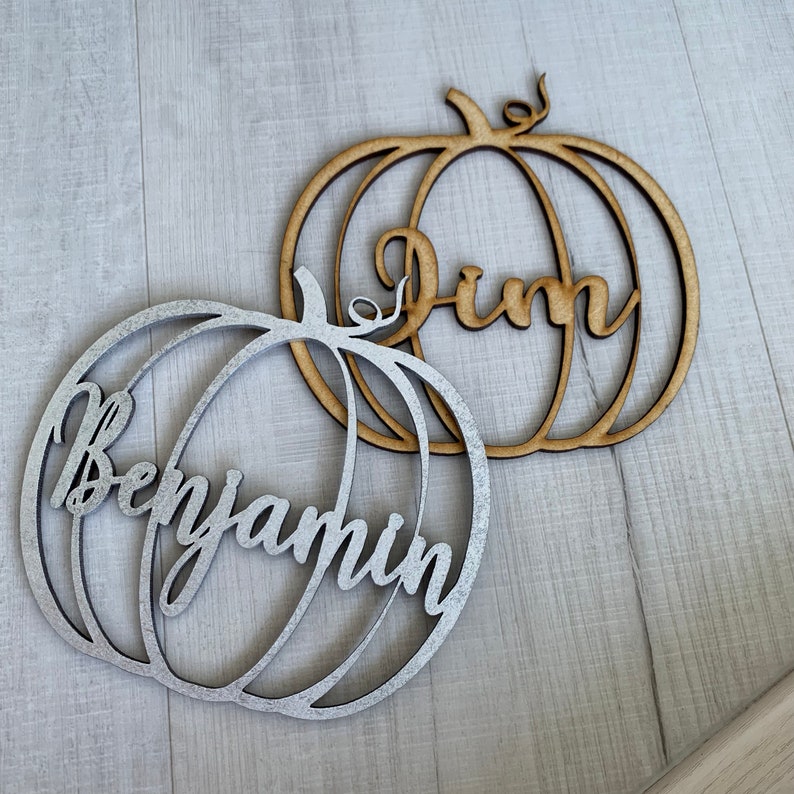 May include: Two wooden pumpkin ornaments with names written on them. The pumpkin on the left is silver and says "Benjamin". The pumpkin on the right is brown and says "Jim".