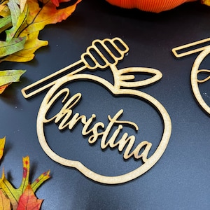 May include: Wooden apple-shaped ornament with a honey dipper on top. The ornament is personalized with the name "Christina".