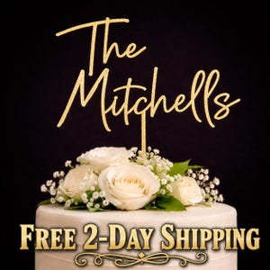 May include: A gold glitter cake topper featuring the text The Mitchells in a cursive script font. The topper is positioned above a white cake decorated with a cluster of white roses and baby's breath flowers against a solid black background.