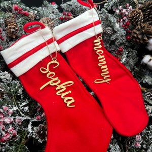 May include: Two red Christmas stockings with white cuffs and gold personalized ornaments. The stockings are decorated with a red and white stripe. The ornaments say "Mommy" and "Sophia".