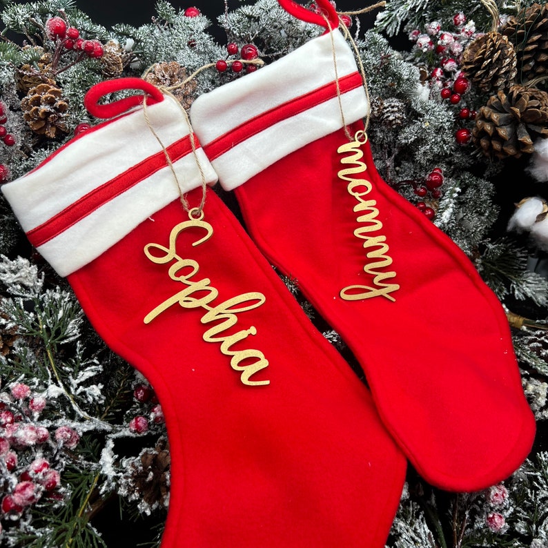 May include: Two red Christmas stockings with white cuffs and gold personalized ornaments. The stockings are decorated with a red and white stripe. The ornaments say "Mommy" and "Sophia".