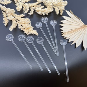 May include: Seven clear acrylic drink stirrers with round tops. The tops are engraved with the names "the Smiths" and "RA 08.08.22".