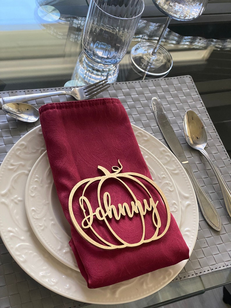 May include: A wooden pumpkin-shaped place card with the name "Johnny" written in cursive. The place card is on a red napkin, which is on a white plate.