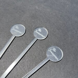 May include: Three silver cocktail stirrers with round, clear tops. Each top has the text "The Jouissance" engraved on it.