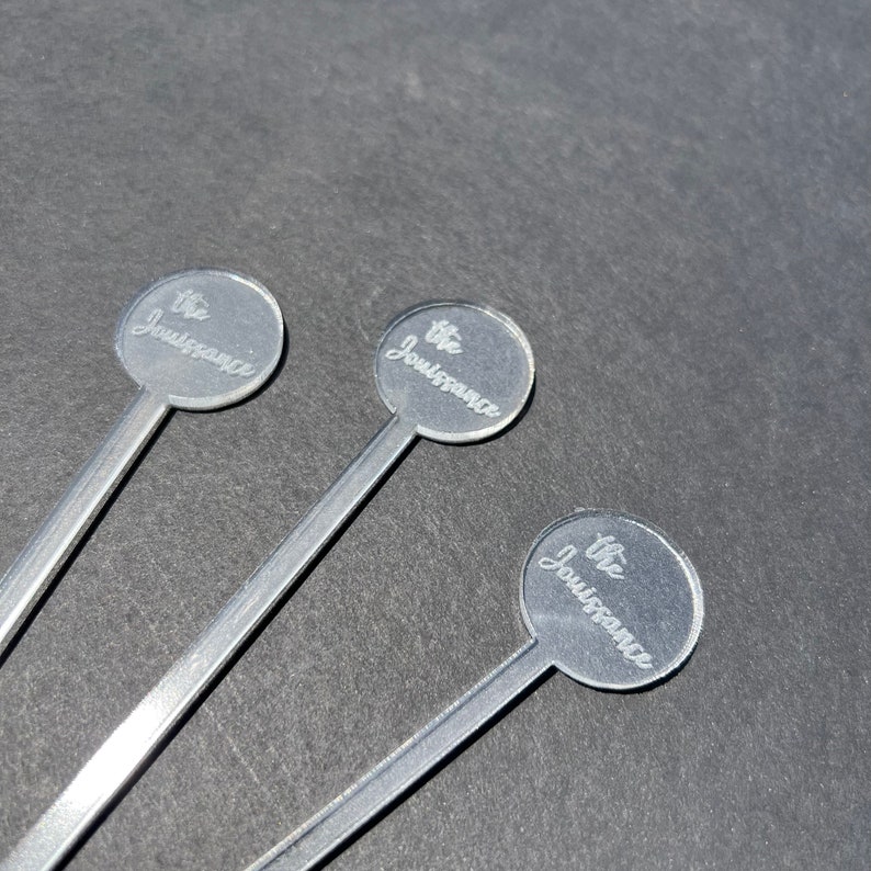 May include: Three silver cocktail stirrers with round, clear tops. Each top has the text "The Jouissance" engraved on it.