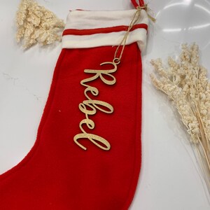 May include: Red Christmas stocking with a white cuff and a gold wooden ornament with the name "Rebel" written on it.