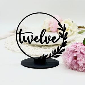 May include: Black table number with a circular frame and the word "twelve" written in a script font. The frame is decorated with a leafy design.