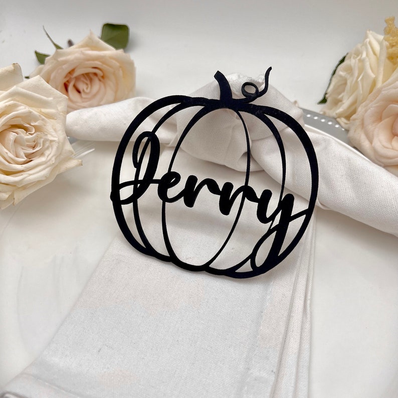 May include: Black pumpkin-shaped napkin ring with the name "Jenny" cut out. The ring is on a white napkin.