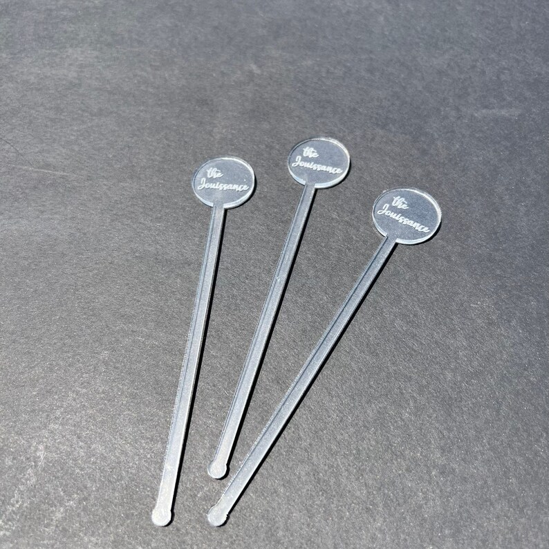 May include: Three clear acrylic cocktail stirrers with a round, silver-colored top. Each top has the text "The Jouissance" engraved on it.