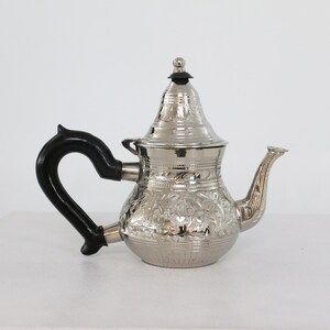 May include: A silver teapot with an ornate floral design and a black handle. The teapot has a lid with a small knob on top.