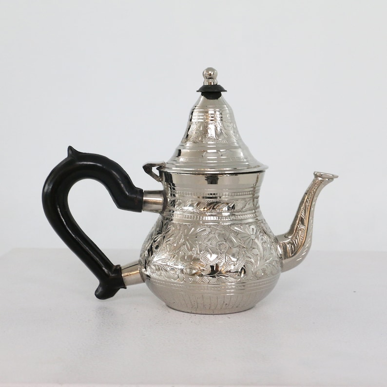 May include: A silver teapot with an ornate floral design and a black handle. The teapot has a lid with a small knob on top.