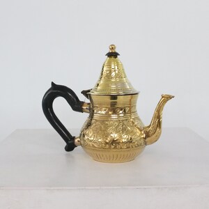 May include: A gold teapot with an ornate floral design and a black handle. The teapot has a domed lid with a finial.