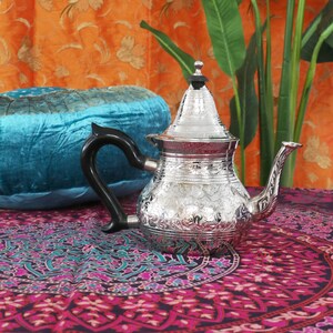 May include: A silver teapot with an ornate floral design sits on a colorful patterned fabric. The teapot has a round body and a tall, pointed lid. The spout is curved and the handle is black.