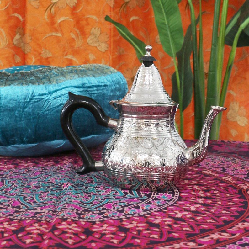 May include: A silver teapot with an ornate floral design sits on a colorful patterned fabric. The teapot has a round body and a tall, pointed lid. The spout is curved and the handle is black.