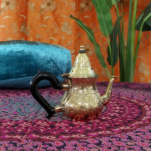 May include: A gold-coloured metal teapot with an ornate design sits on a colourful patterned fabric. The teapot has a black handle and a spout.