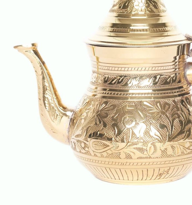 May include: A gold-colored metal teapot with an ornate floral design. The teapot has a lid and a spout.