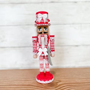 May include: A red and white nutcracker figurine with a white bow and a silver sword. The nutcracker is wearing a red hat with white trim and a white lace collar.