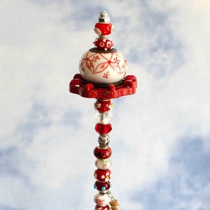 May include: A whimsical red and white ceramic bead garland with a small glass vial charm. The garland is displayed in a clear glass bowl filled with colorful glass stones.