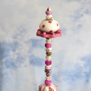 May include: A pink and white ceramic knob with red polka dots sits atop a tall, decorative beaded tower. The tower is made of various beads, including pink, white, and purple glass beads, and floral patterned beads. The tower is placed in a clear glass bowl filled with green and blue glass stones.