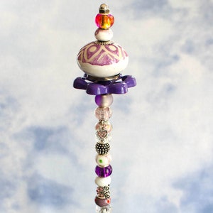 May include: A decorative beaded ornament with a variety of colorful beads, including purple, pink, and silver. The ornament has a white and purple patterned top and a purple flower-shaped base. The ornament is displayed in a glass bowl.