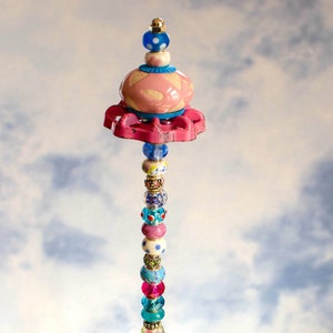 May include: A colorful beaded garden stake with a pink and white ceramic top and a pink and white ceramic bottom. The stake is standing in a glass bowl filled with green and blue glass stones.