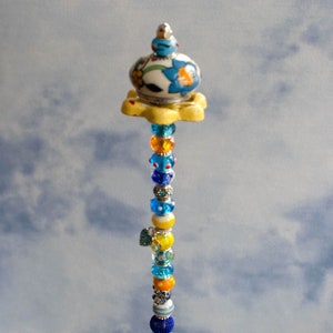 May include: A decorative beaded stem with a floral ceramic top and base, featuring a variety of colorful beads in shades of blue, yellow, orange, and silver. The stem is displayed in a clear glass bowl filled with decorative glass stones.