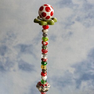 May include: A tall, decorative beaded stick with a red and white polka dot ceramic ball on top. The stick is adorned with colorful beads, including red, green, white, and silver. The stick is standing in a clear glass bowl filled with blue and green glass stones.