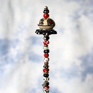 May include: A tall, decorative beaded stem with a black and white ceramic top, red and black beads, and a silver charm. The stem is standing in a clear glass bowl filled with blue and green glass beads.