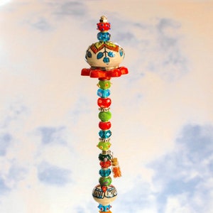 May include: A colorful beaded tower with a ceramic top and a small glass vial hanging from it. The tower is standing in a clear glass bowl filled with blue and green glass stones.