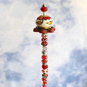 May include: A red and white ceramic bead garland with a decorative ceramic top. The garland is hanging from a clear glass bowl filled with green and blue glass beads.