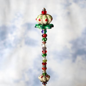 May include: A decorative beaded wand with a white ceramic top featuring a floral design. The wand is adorned with red, green, and white beads and is displayed in a clear glass bowl filled with blue and green glass stones.