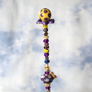 May include: A purple and yellow beaded garland with a purple and white polka dot mushroom charm at the top and bottom. The garland is in a clear glass vase filled with green and blue glass beads.