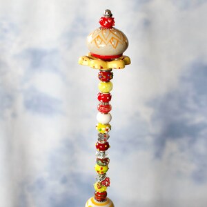 May include: A tall, decorative beaded string with a white and yellow ceramic top and bottom. The beads are red, yellow, white, and clear glass. The string is in a clear glass bowl with blue and green glass stones.