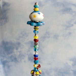 May include: A colorful beaded garden stake with a blue and white ceramic top. The stake is topped with a blue and white ceramic knob with yellow stars. The stake is surrounded by green glass beads.
