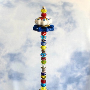 May include: A tall, decorative beaded object with a variety of colorful beads, ceramic elements, and a blue and white floral design. The object is displayed in a clear glass bowl filled with colorful glass stones.