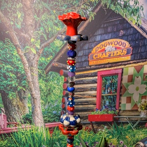 May include: A colorful garden stake with a red flower on top, blue and white beads, and a blue and white ceramic knob. The stake is in front of a wooden building with a sign that says "Dogwood Crafters, A Craft Cooperative".