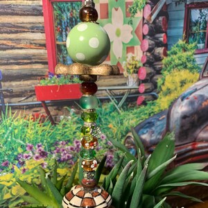 May include: A whimsical garden stake made from repurposed materials. It features a green ceramic ball with white polka dots, a brown and white ceramic knob, and a variety of colorful beads and charms. The stake is surrounded by green succulents.