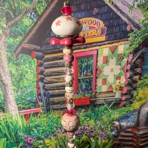 May include: A pink and white ceramic garden stake with a star design and beads. The stake is decorated with pink, white, and clear beads. The stake is in front of a wooden building with a sign that says "Wood Crafters".