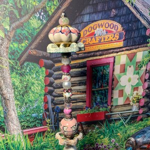 May include: A pink and white ceramic garden stake with a whimsical design. The stake is decorated with beads, charms, and a small ceramic bird. The stake is in front of a wooden building with a sign that reads "Dogwood Crafters Art Cooperative".