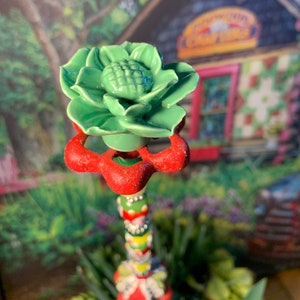 May include: A green ceramic flower with a red and white base. The flower is on a colorful pole with beads and other decorations. The pole is in front of a blurred background of a house with a sign that says "Country Charm".