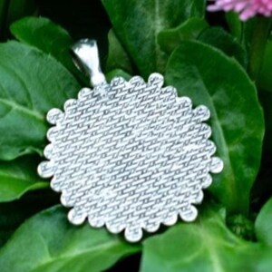 May include: A silver-colored pendant with a scalloped edge, resting on a bed of green leaves. The pendant has a textured surface and a small loop for a chain. The background features more green foliage and a hint of a pink flower.