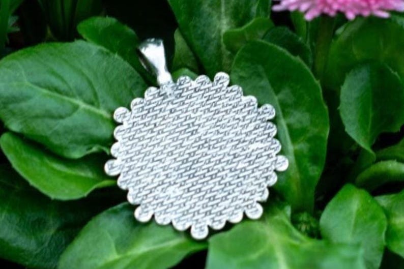 May include: A silver-colored pendant with a scalloped edge, resting on a bed of green leaves. The pendant has a textured surface and a small loop for a chain. The background features more green foliage and a hint of a pink flower.