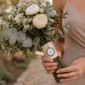 May include: A bouquet of white flowers, including roses and daisies, with green eucalyptus leaves. The stems are wrapped with a cream-colored ribbon and a silver pendant. The bouquet is held by a person wearing a beige dress.