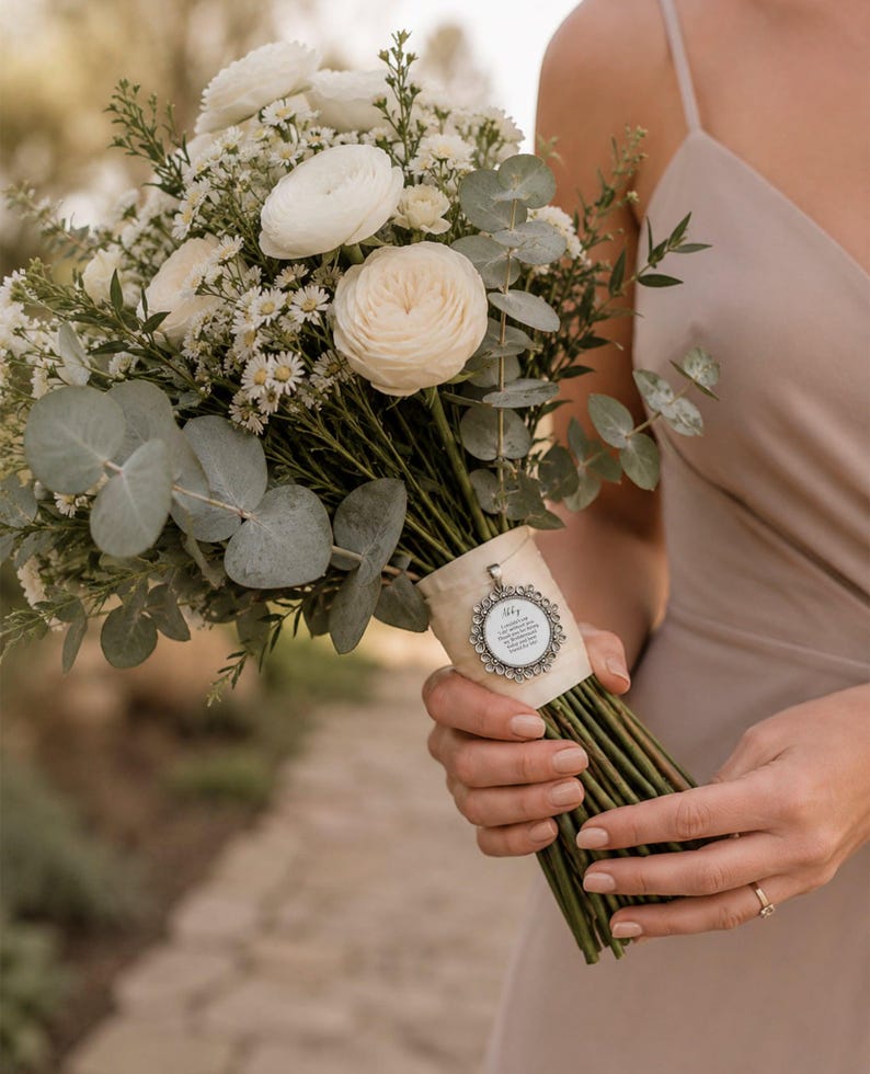 May include: A bouquet of white flowers, including roses and daisies, with green eucalyptus leaves. The stems are wrapped with a cream-colored ribbon and a silver pendant. The bouquet is held by a person wearing a beige dress.