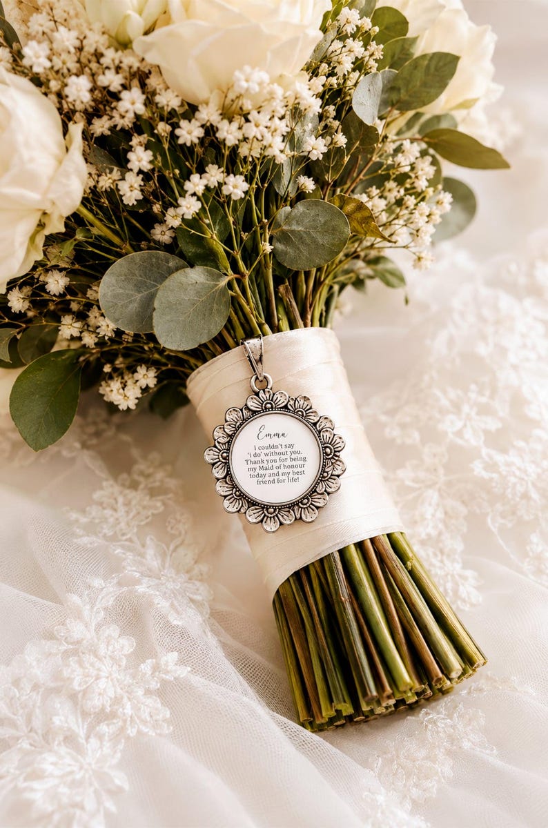 May include: A bridal bouquet featuring white roses, baby's breath, and eucalyptus leaves. The stems are wrapped with a cream-colored ribbon, and a silver pendant with the name "Emma" and a message hangs from the ribbon.